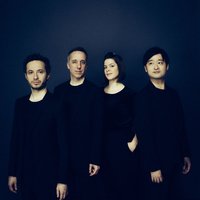 Four individuals in dark clothing stand against a dark background. They look seriously into the camera.