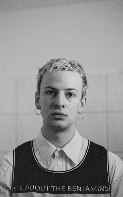Ein schwarz-weißes Bild von einer Person in einem Hemd mit einem Oberteil darüber, kurzen Haaren und großen Ohrringen. Die Person blickt in die Kamera mit einem ernsten Gesichtsausdruck.