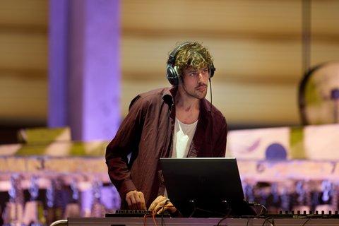 Ein DJ steht an einem Mischpult mit Laptop, trägt Kopfhörer und ist in ein musikalisches Set vertieft.