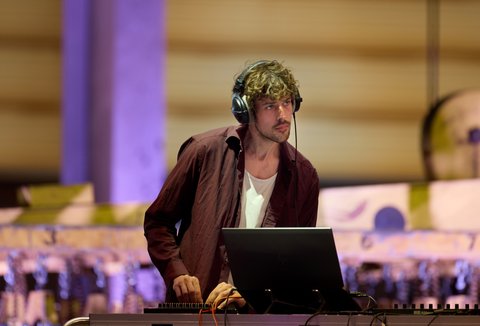 Ein DJ steht an einem Mischpult mit Laptop, trägt Kopfhörer und ist in ein musikalisches Set vertieft.