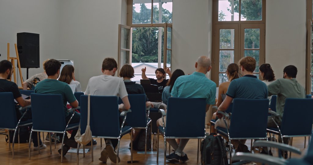 Eine Gruppe von Menschen von hinten. Vor ihnen steht ein Mann der dirigiert.