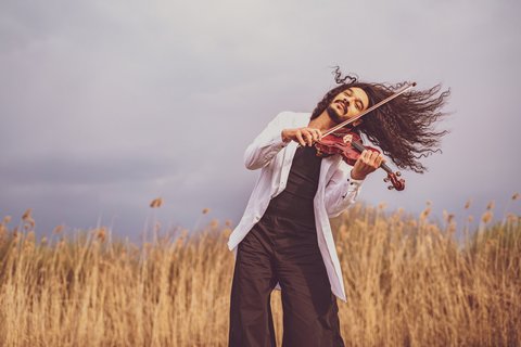A person passionately plays the violin in a field of tall grass under a cloudy sky.