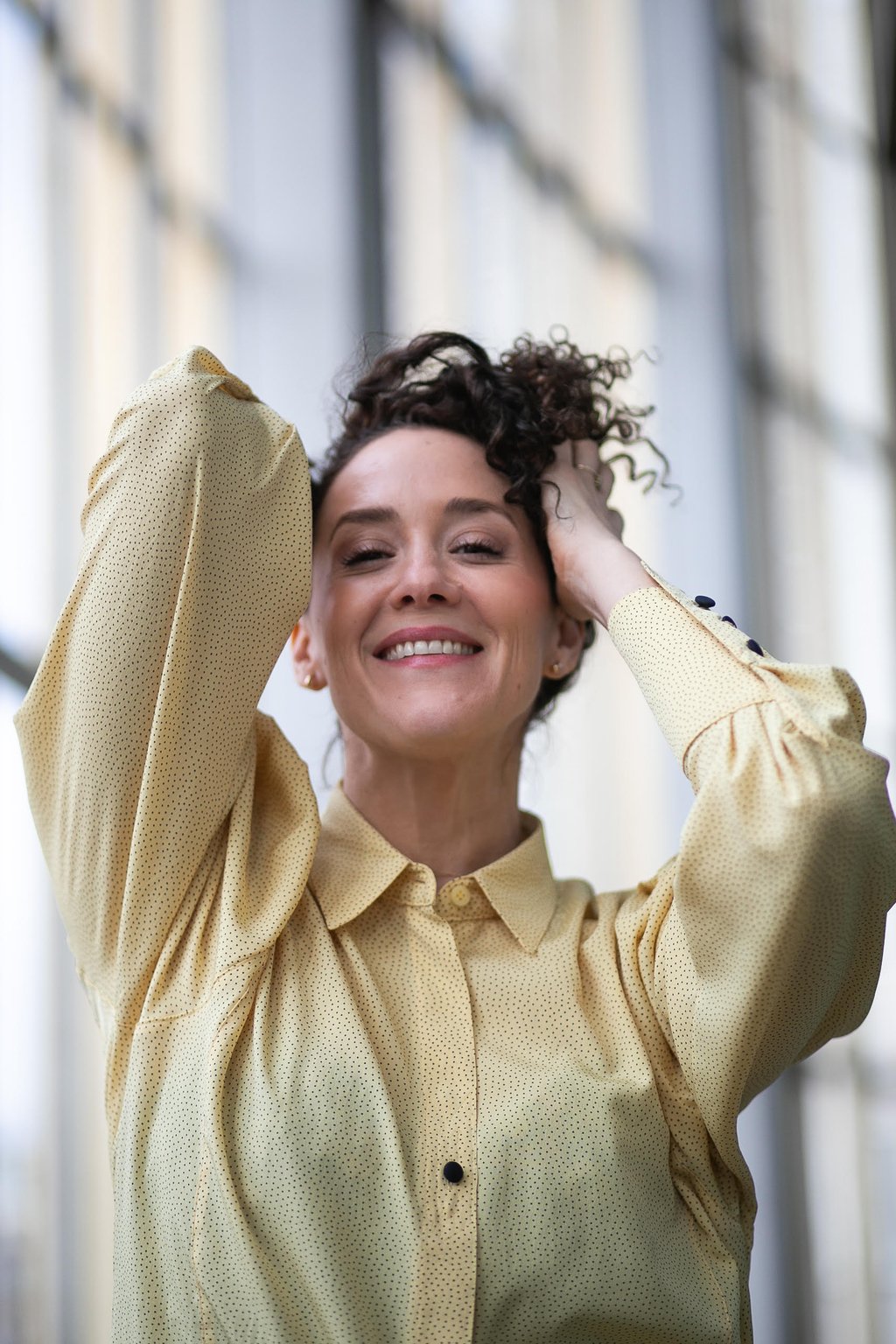 Eine Frau mit schwarzen Locken, sie hat ihre Hände in ihren Haaren. Sie trägt eine gelbe Bluse und lächelt.