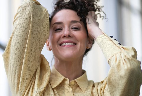 Eine Frau mit schwarzen Locken, sie hat ihre Hände in ihren Haaren. Sie trägt eine gelbe Bluse und lächelt.