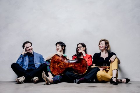 Eine Gruppe von vier Musikern sitzt lachend auf dem Boden vor einer weißen Wand. Einer hält ein Streichinstrument.