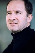 Ein Mann im Porträt, er schaut ernst an der Kamera vorbei. Er trägt einen schwarzen Rollkragenpullover. Der Hintergrund ist türkis-grau.