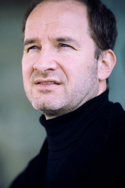 Ein Mann im Porträt, er schaut ernst an der Kamera vorbei. Er trägt einen schwarzen Rollkragenpullover. Der Hintergrund ist türkis-grau. © anna.s