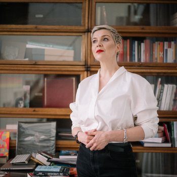 Eine Frau steht vor einem Bücherregal. Sie blickt von der Kamera weg. Ihre Haare sind kurz und sie trägt eine helle Bluse.