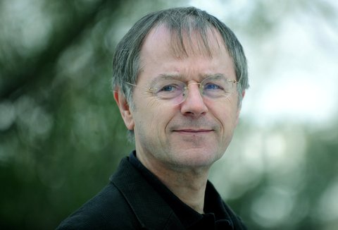 Portrait of a man outdoors in a black suit, with a blurred background of trees or shrubs.