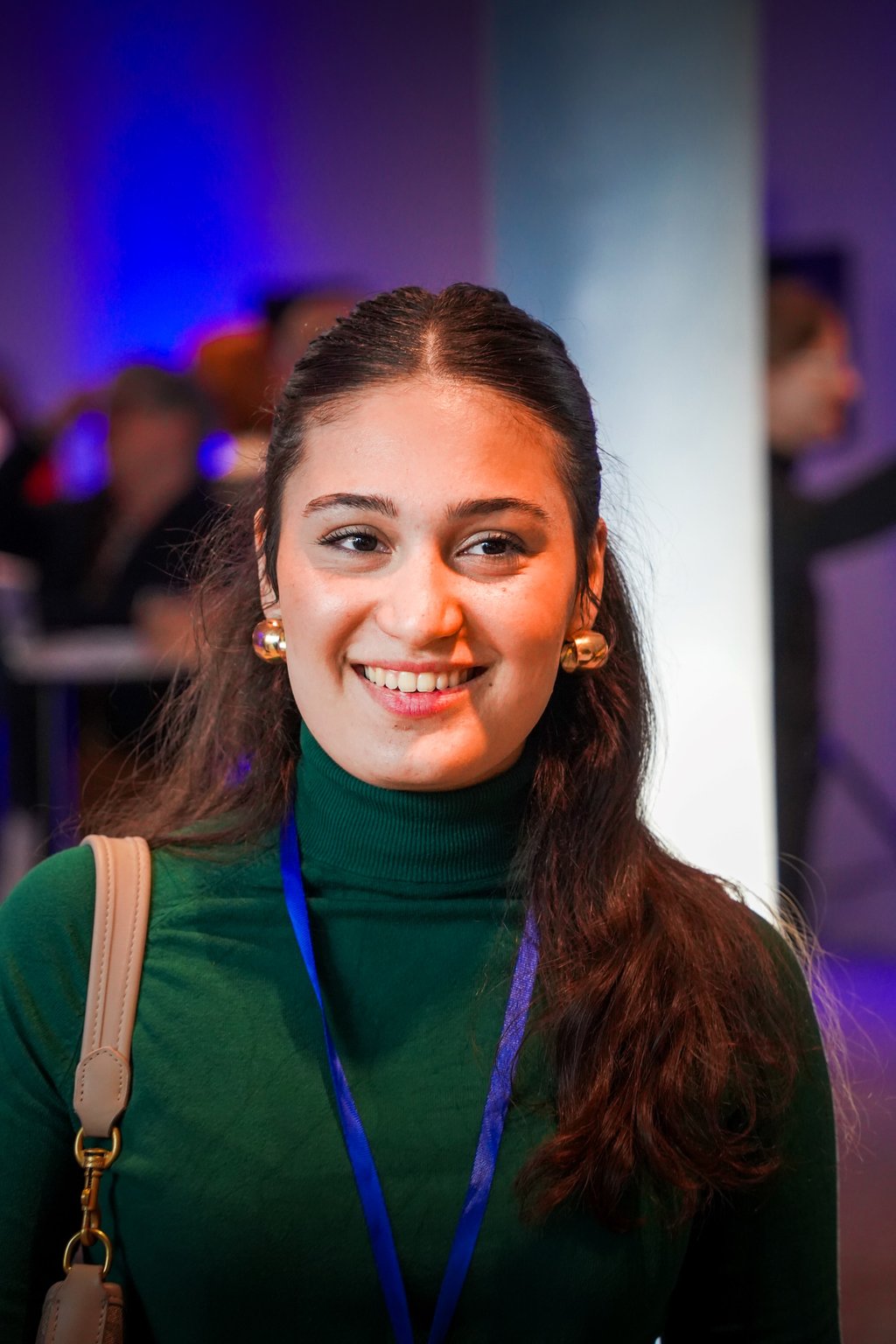 A woman in a green turtleneck smiles, wearing a shoulder bag and a blue lanyard. The background is blurred with blue lights.
