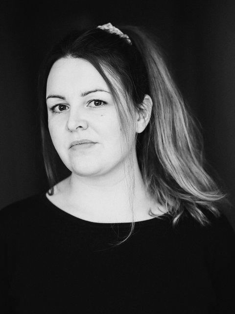A woman is standing in front of a dark background and looking into the camera.