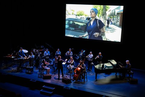 Eine Gruppe von Musikern spielt auf der Bühne vor einem großen Bildschirm, der das Bild einer Frau zeigt.