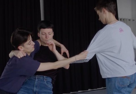 Three dancers are intertwined in a pose, practicing in a studio with a black curtain. They wear casual clothing.