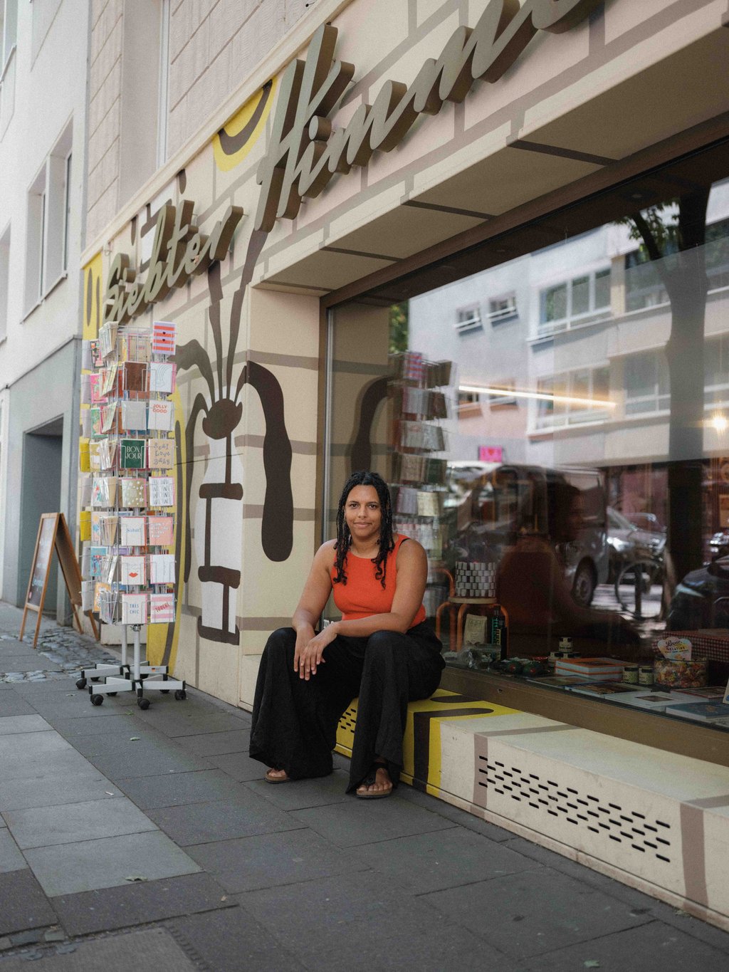 Eine Frau in einem roten Oberteil sitzt vor einer Buchhandlung. Sie posiert für die Kamera und lächelt.