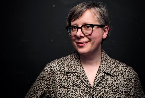 A woman is standing in front of a dark background. She is wearing a patterned blouse. She is looking into the camera and smiling.