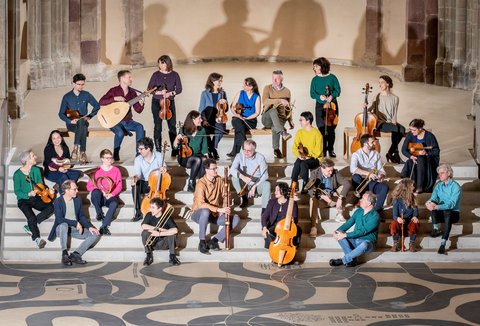 A group of musicians in colourful clothing is sitting on different steps of a broad staircase. Everybody is holding their instruments and talking animatedly.