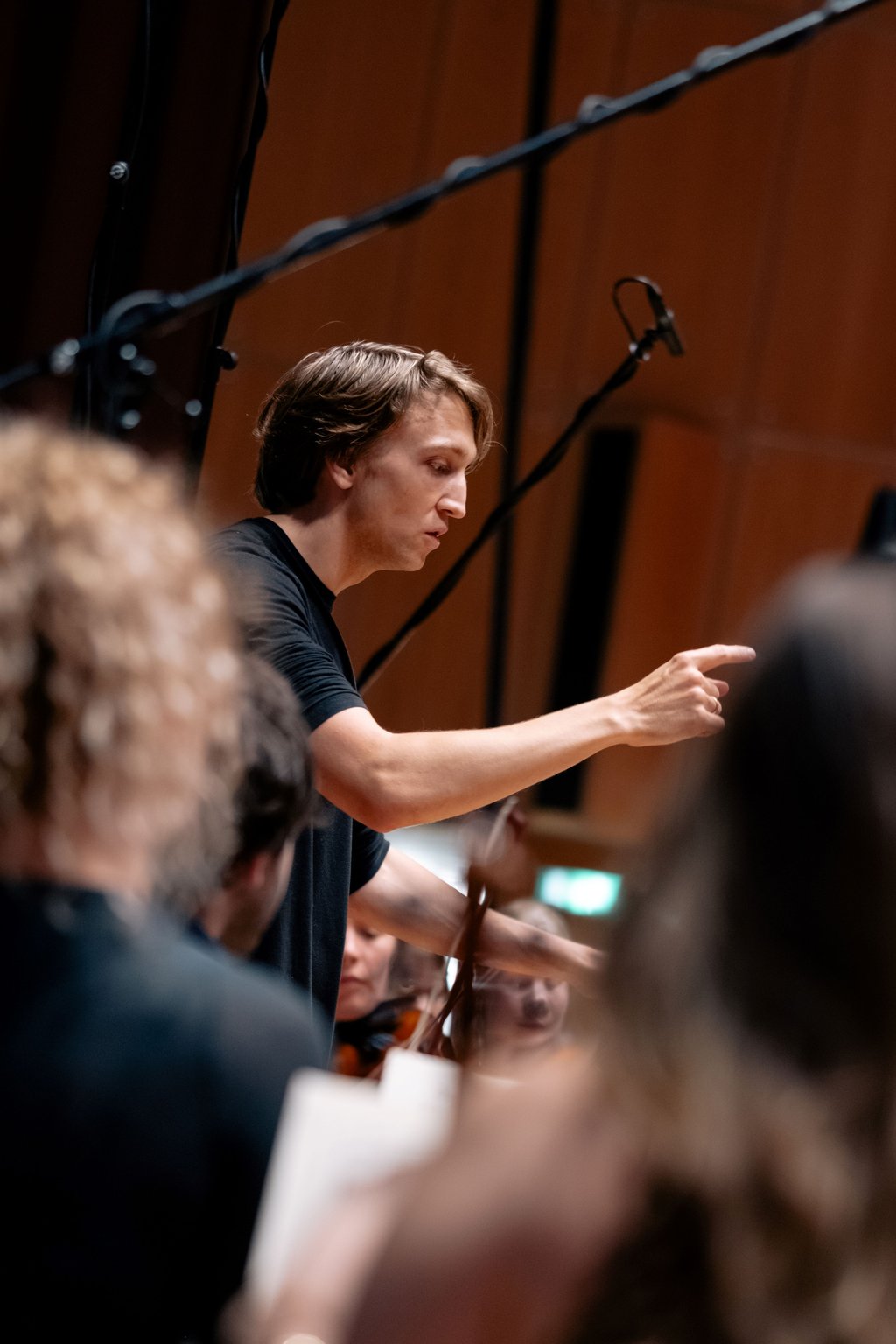 Ein Dirigent in schwarzem T-Shirt dirigiert ein Orchester. Der Fokus liegt auf seiner energischen Gestik und Aufmerksamkeit.