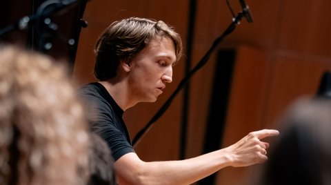 Ein Dirigent in schwarzem T-Shirt dirigiert ein Orchester. Der Fokus liegt auf seiner energischen Gestik und Aufmerksamkeit.