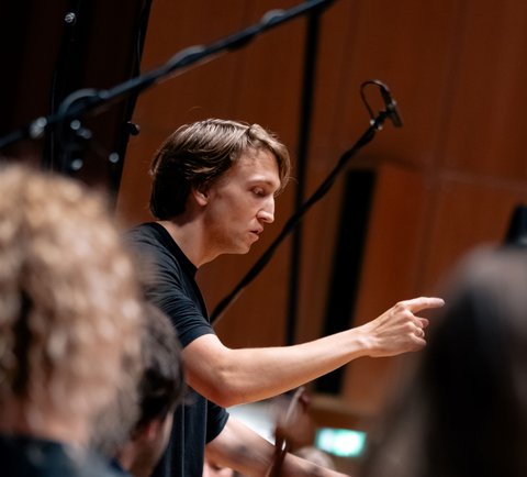 Ein Dirigent in schwarzem T-Shirt dirigiert ein Orchester. Der Fokus liegt auf seiner energischen Gestik und Aufmerksamkeit.