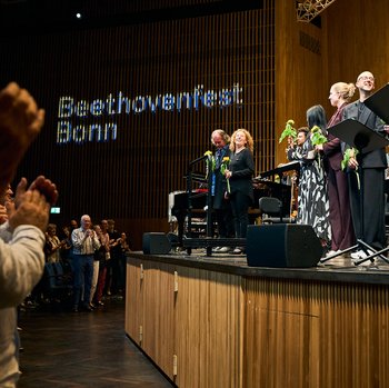 Menschen auf der Bühne und im Publikum beim Beethovenfest Bonn. Applaus erfüllt den Raum, während Künstler Blumen erhalten.