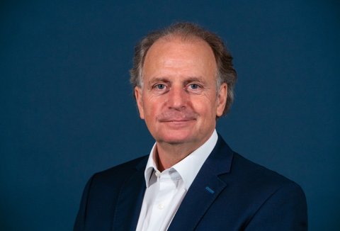 Ein Mann im Anzug posiert für ein Portrait vor einem dunkelblauen Hintergrund. Er lächelt in die Kamera.