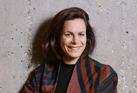 Eine Frau in einer gemusterten Bluse posiert mit verschränkten Armen. Sie blickt in die Kamera und lächelt.