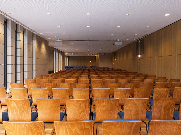 An empty conference room with numerous rows of chairs, well-lit by ceiling lights and large windows on the left side.