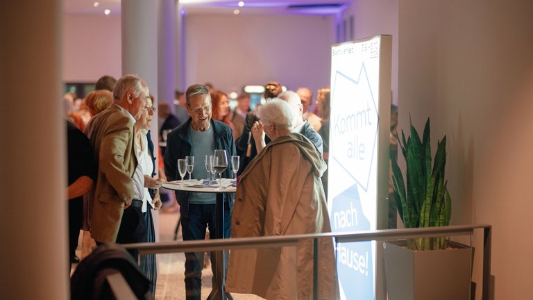 People standing in an elegantly lit gallery or event space, chatting and enjoying drinks. Plants and signage are visible.