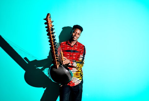 Ein Musiker in einem bunten Hemd spielt eine Kora vor einem hellblauen Hintergrund. Sein Gesichtsausdruck ist konzentriert.
