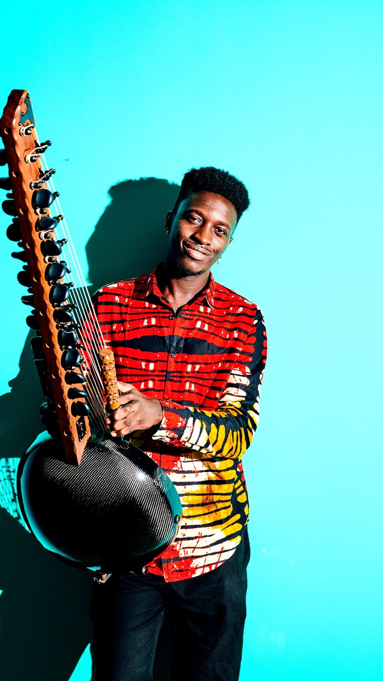 Ein Musiker in einem bunten Hemd spielt eine Kora vor einem hellblauen Hintergrund. Sein Gesichtsausdruck ist konzentriert.