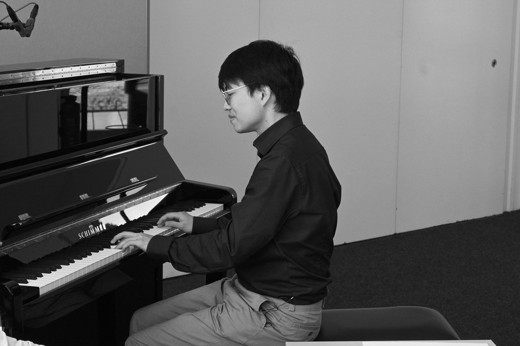 Schwarz-Weiß-Foto eines Mannes, der konzentriert Klavier spielt. Der Raum ist schlicht, mit einem aufrechten Klavier.