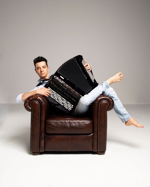 Ein Mann posiert mit seinem Akkordeon vor einem hellen Hintergrund. Er streckt sich barfuß in einem Sessel und blickt in die Kamera. © Pigini Accordions