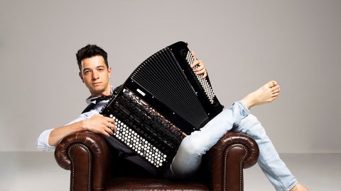 A man is posing with his accordion before a light background. He is lounging barefoot in a chair and looking into the camera.