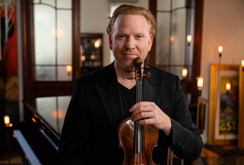 A man holds a violin in an elegant room, with mirrors and candlelight in the background, creating a warm ambiance.