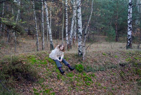 A woman is sitting in the forest. Her legs are stretched out on the ground before her. She is wearing a light-coloured jacket. She is looking into the camera.