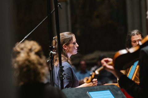 Ein Orchester probt, mit Musikern, die konzentriert spielen. Im Vordergrund sind Noten und ein Mikrofon zu sehen.