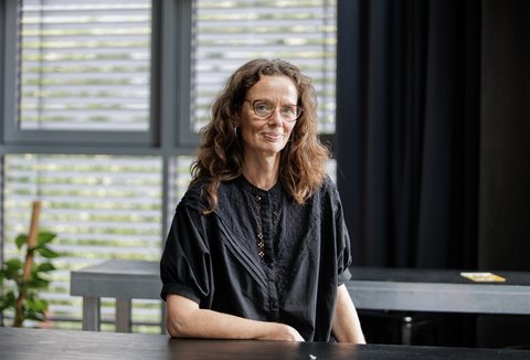 A woman with long brown hair, glasses and a black top is standing in a well-lit room. She is looking into the camera.