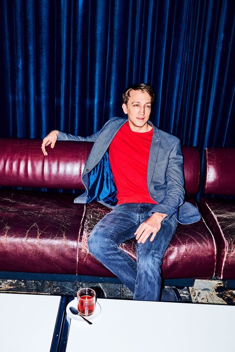 Ein Mann in blauem Blazer und rotem T-Shirt sitzt entspannt auf einem roten Sofa mit einem Drink auf dem Tisch.