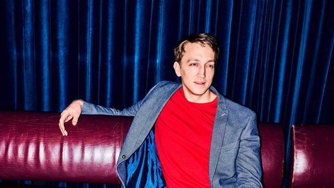 A man in a blue blazer and red T-shirt sits relaxed on a red sofa with a drink on the table.  © Patrick Essex