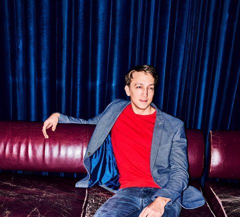 A man in a blue blazer and red T-shirt sits relaxed on a red sofa with a drink on the table.