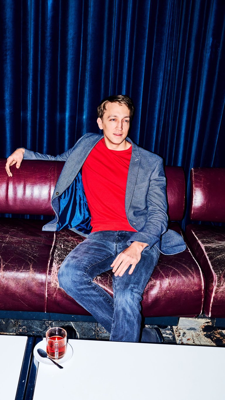 Ein Mann in blauem Blazer und rotem T-Shirt sitzt entspannt auf einem roten Sofa mit einem Drink auf dem Tisch.