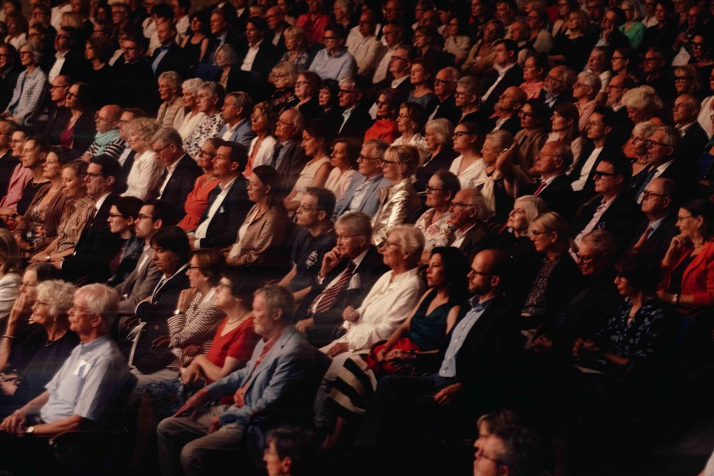 Ein dicht gefülltes Auditorium mit Menschen, die aufmerksam einem Event folgen. Unterschiedliche Altersgruppen sind präsent, in formeller Kleidung.