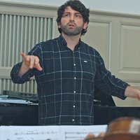 A conductor leads a rehearsal in a music room. A pianist plays in the background. Music sheets are in the foreground.