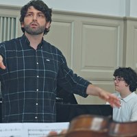 A conductor leads a rehearsal in a music room. A pianist plays in the background. Music sheets are in the foreground.