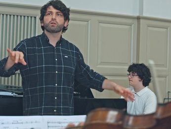 Ein Dirigent leitet eine Probe in einem Musikraum. Ein Pianist spielt im Hintergrund. Partituren sind im Vordergrund.