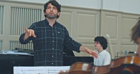 Ein Dirigent leitet eine Probe in einem Musikraum. Ein Pianist spielt im Hintergrund. Partituren sind im Vordergrund.