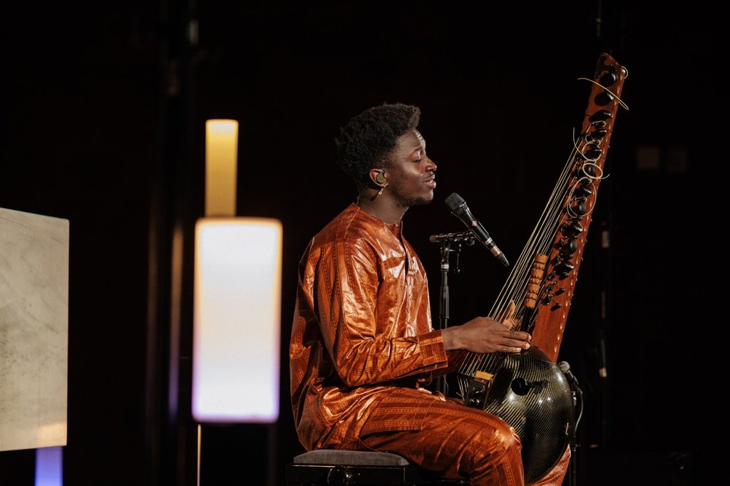 Ein Musiker in einem glänzenden orangenen Outfit spielt auf einer traditionellen westafrikanischen Kora und singt in ein Mikrofon.