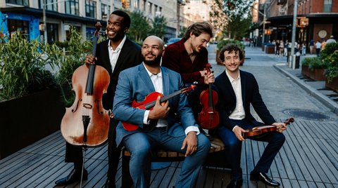 A group of four musicians with classical instruments smiling and posing outdoors on a promenade in a city setting.