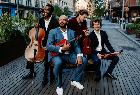 Eine Gruppe von vier Musikern mit klassischen Instrumenten posiert lachend im Freien auf einer Promenade in einer Stadt.