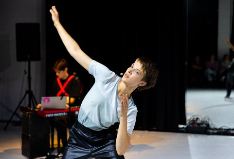 A woman moves in front of a dark background. She is wearing dancer's clothes. Her arms are positioned in a dancing figure.